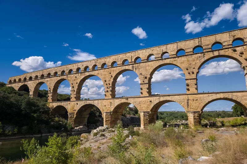 A stone aqueduct bridge with tiers of arches crossing a river under a blue sky.