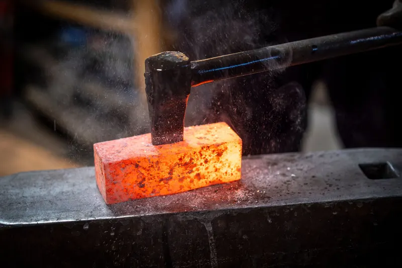 A hammer striking a glowing block of metal resting on an anvil.