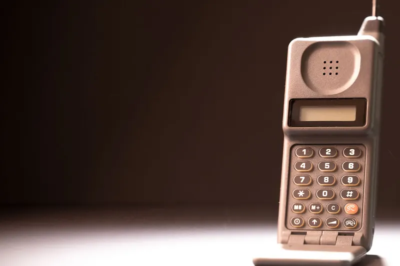 A first-generation portable cell phone with a numeric keypad and a short antenna against a dark background.